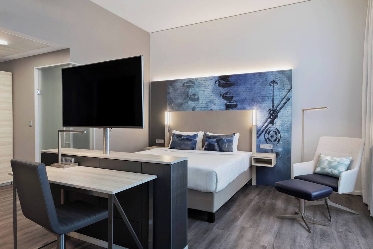 A modern hotel room with a bed, desk, chair, TV, and artwork on the wall.