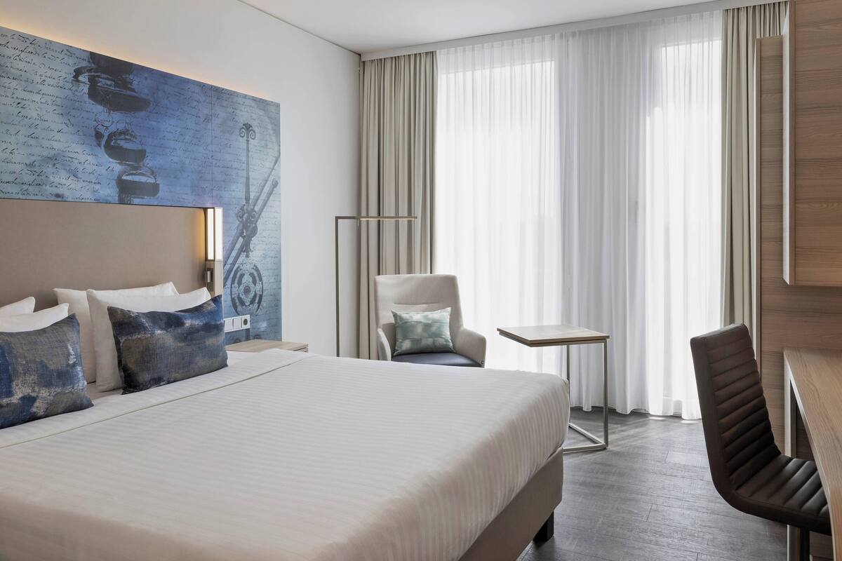 A hotel room with a large bed, a desk, a chair, and a view of the outdoors through sheer curtains.