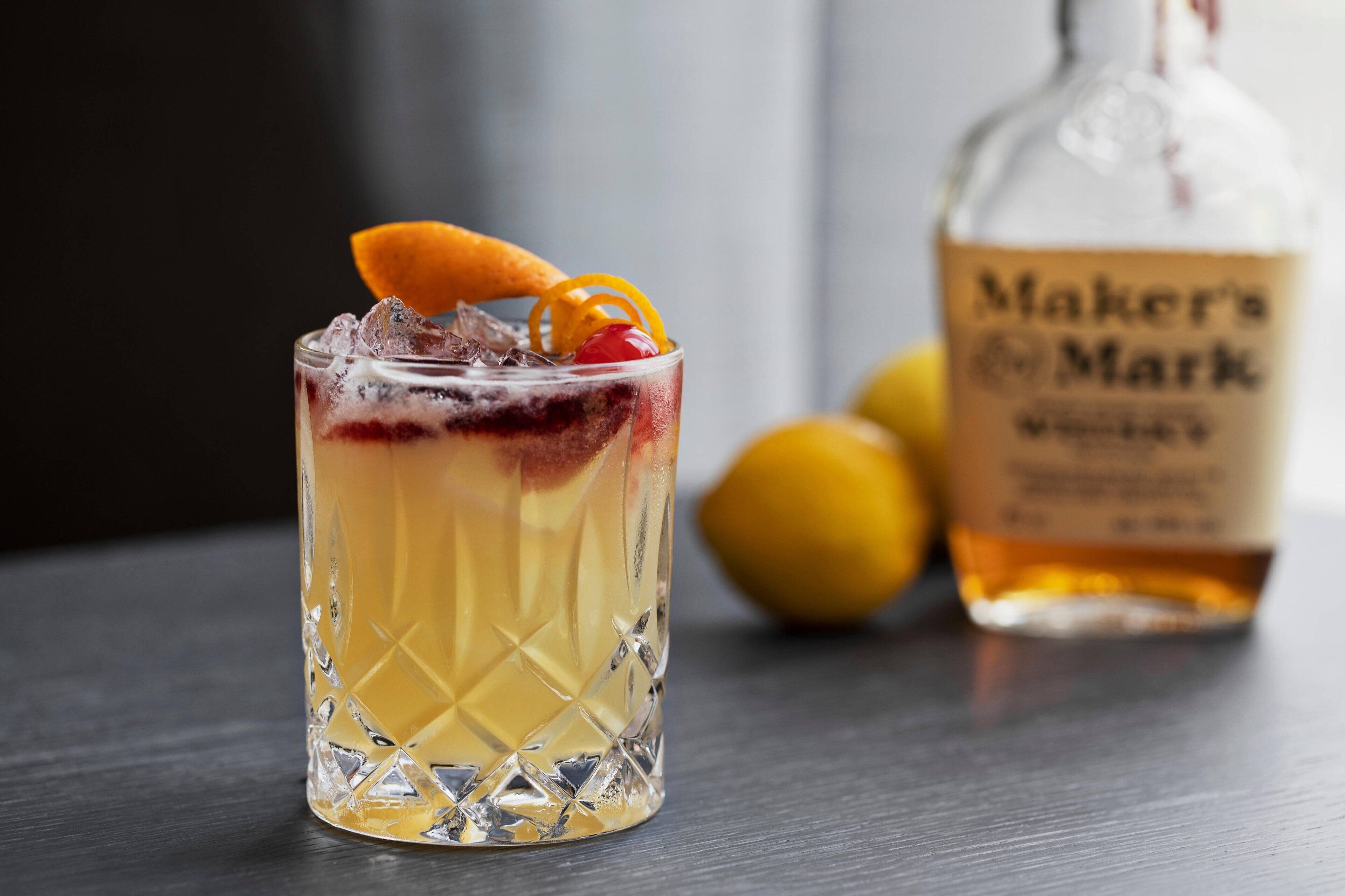 Un verre à cocktail contenant une boisson à plusieurs couches, des glaçons et une cerise enfilée sur une brochette. Une bouteille de whisky Maker's Mark est visible à l'arrière-plan.