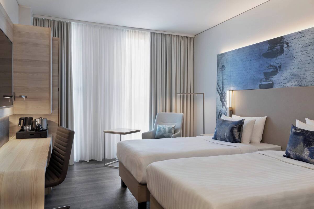 A hotel room with two beds, a desk, a chair, a small table, and a large painting on the wall.