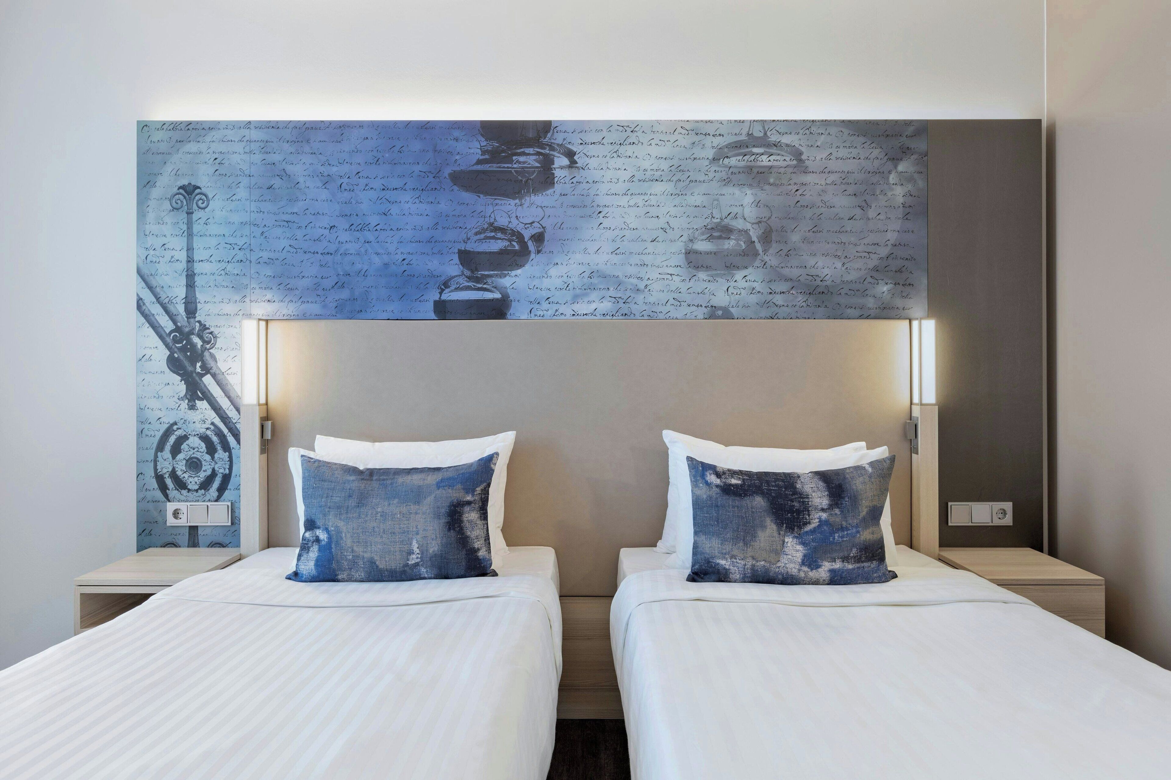 A hotel room with two beds, each with a pillow and a headboard. There is a wall-mounted artwork above the beds.