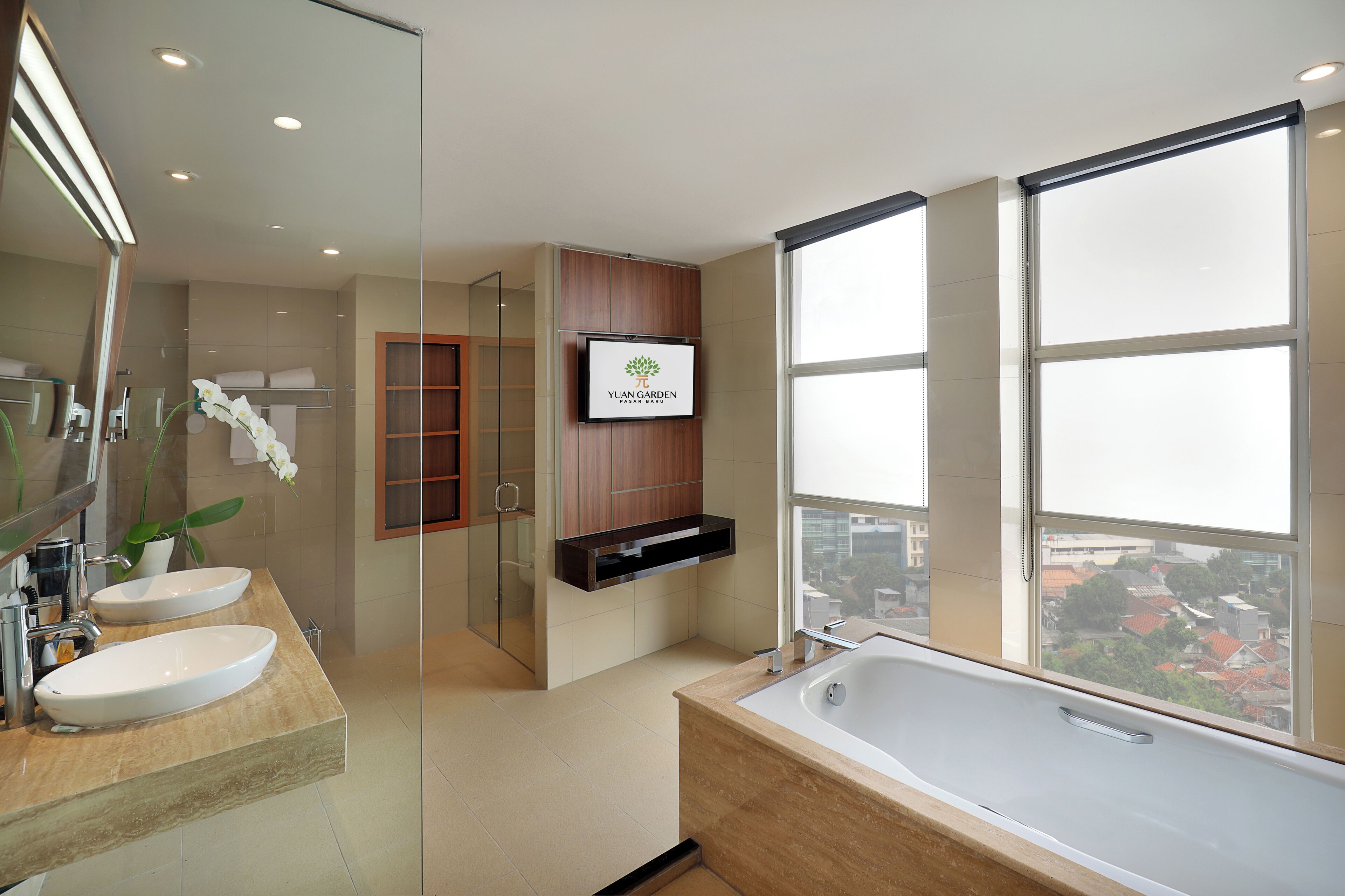 A modern bathroom with two sinks, a large bathtub, a TV mounted on the wall, and a view of the city through large windows.