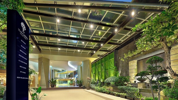Een hotel lobby met glazen plafond, potplanten en een bord waarop voorzieningen staan zoals kamers, een restaurant en een wellnesscentrum.