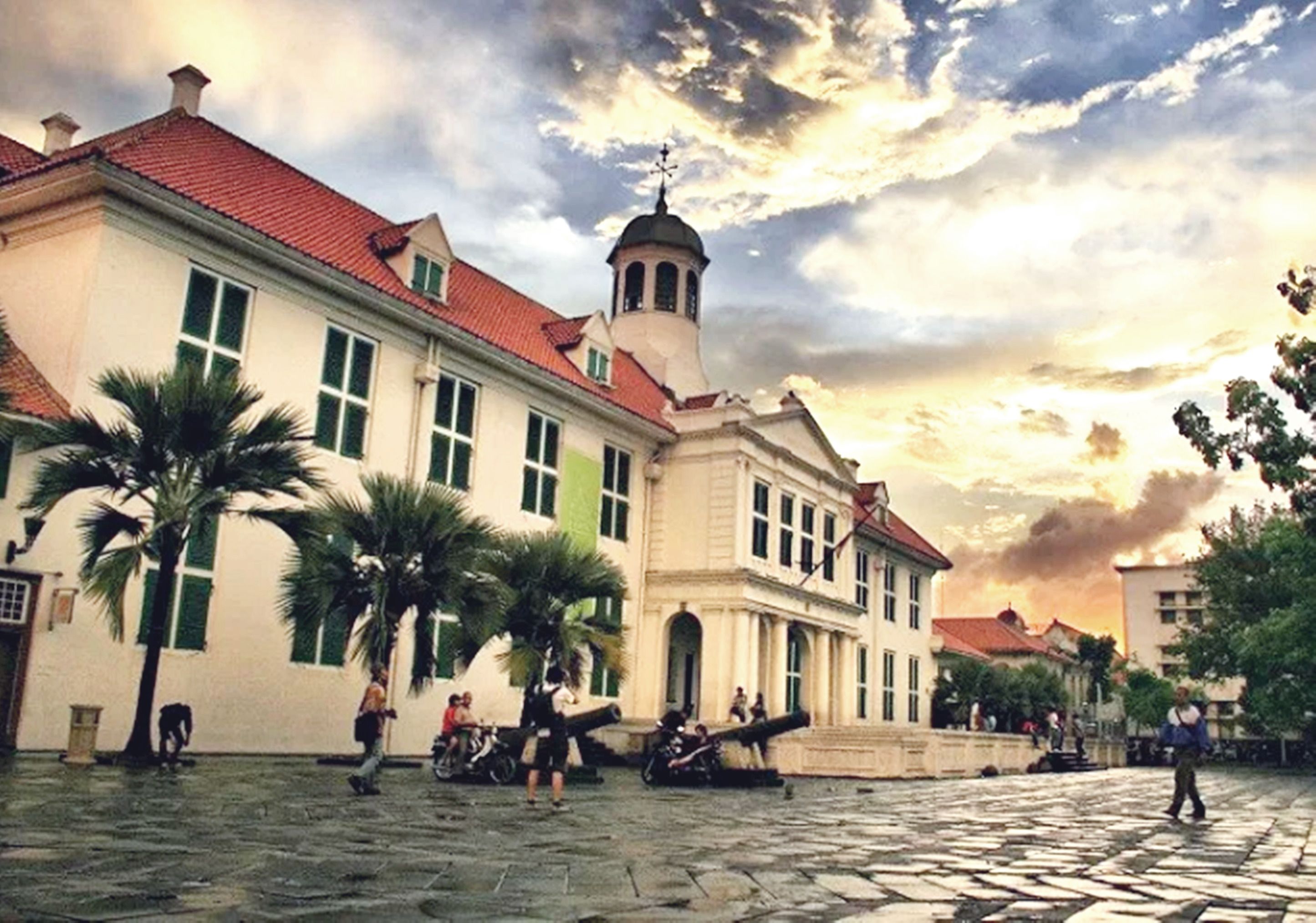 A historic building with a red-tiled roof and a bell tower, surrounded by palm trees and cobblestone paving.