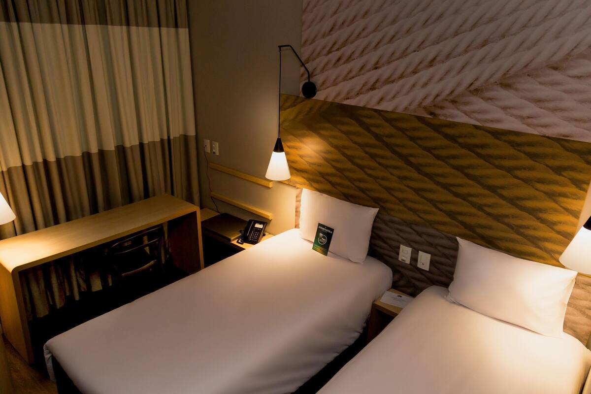 A hotel room with two beds, a desk, a telephone, and a wall-mounted lamp.