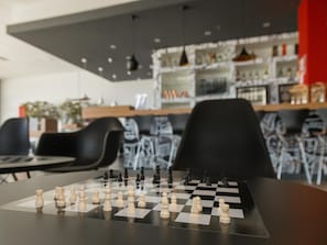 A chessboard with pieces set up on a table in a modern interior space with black chairs and a bar area in the background.