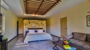 A bedroom with a stone headboard, a bed, a wooden ceiling, a sofa, and a coffee table with books and a fruit basket. - Zornitza Family Estate (Zornitza)