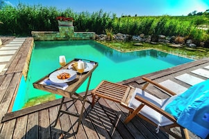 Um deck de madeira com piscina, uma mesa com comida e bebidas, e cadeiras de descanso.