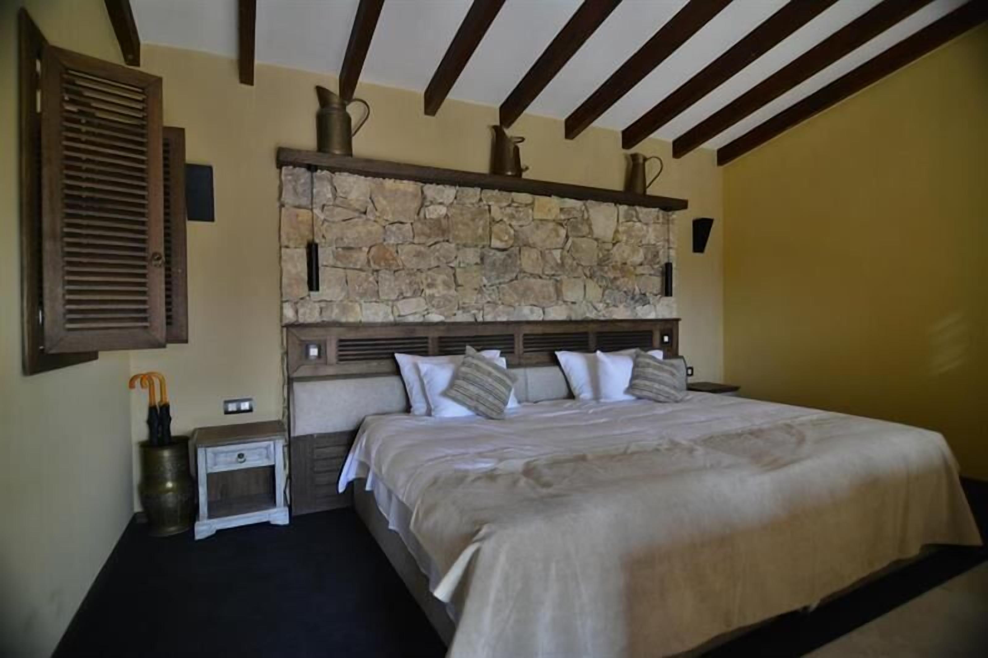 A bedroom with a stone headboard, wooden beams, a bedside table with a lamp, and a bed with white bedding.