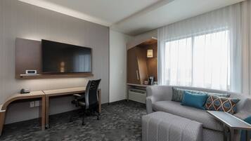 A hotel room with a desk, a flat-screen TV, a sofa, and a large window with curtains.