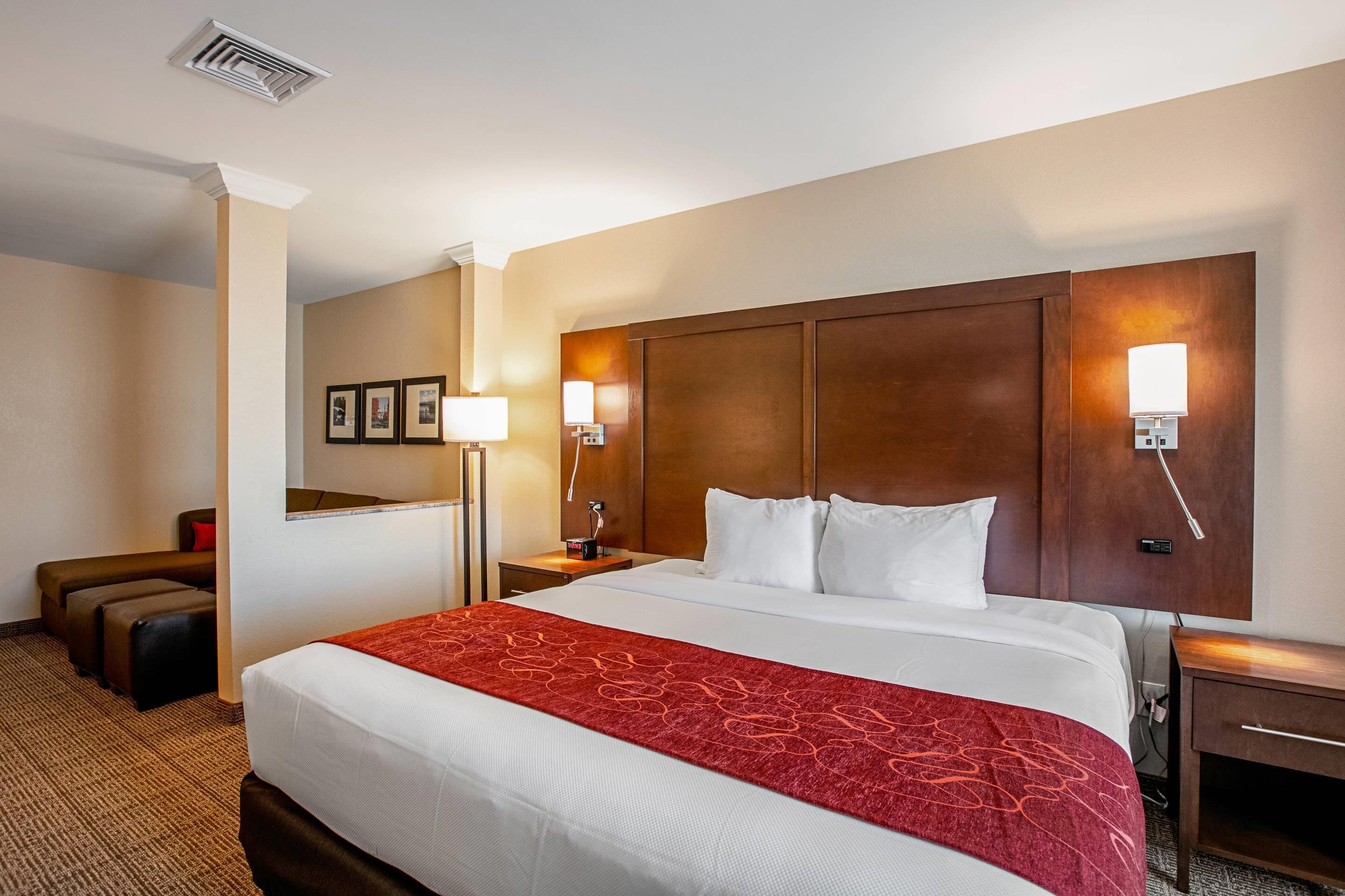A hotel room with a large bed, a wooden headboard, two bedside lamps, a desk, and a seating area with a sofa and ottoman.
