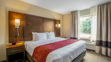 A hotel room with a large bed, two bedside tables, a nightstand, a red throw blanket, a headboard, two wall-mounted lamps, and a window with curtains.
