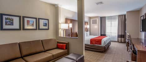 A hotel room with a sofa, ottoman, a bed with a red blanket, a desk, a TV, and framed pictures on the wall.