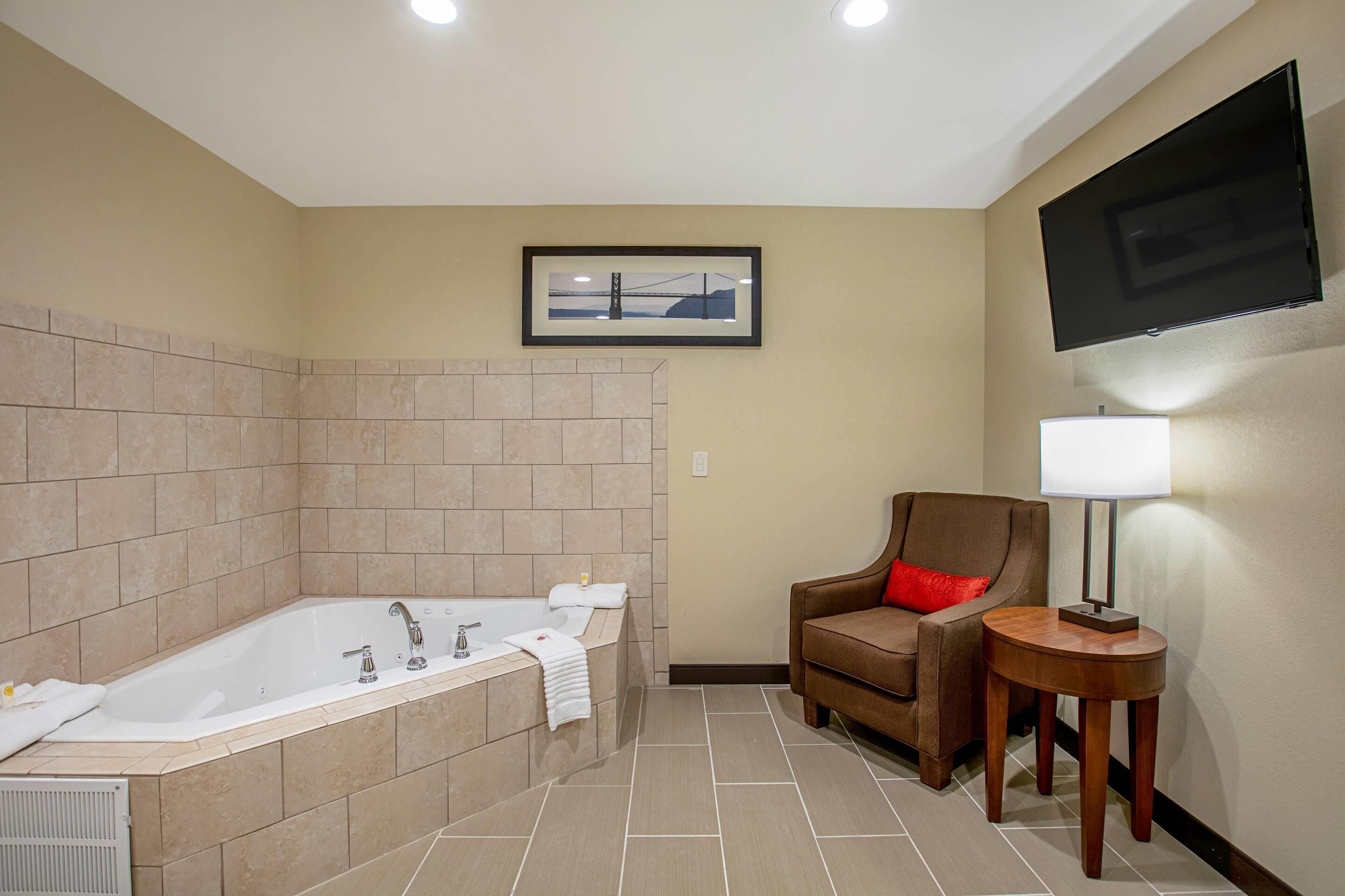 A bathroom with a large bathtub, a chair, a small table with a lamp, and a framed picture on the wall.