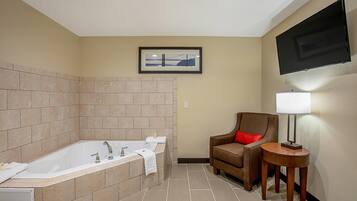 A bathroom with a large bathtub, a chair, a small table with a lamp, and a framed picture on the wall.