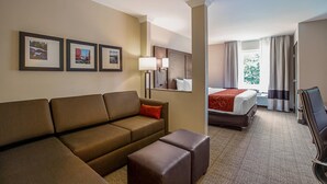 Habitación de hotel con sofá, reposapiés, cama con manta roja, una silla y cuadros en la pared.