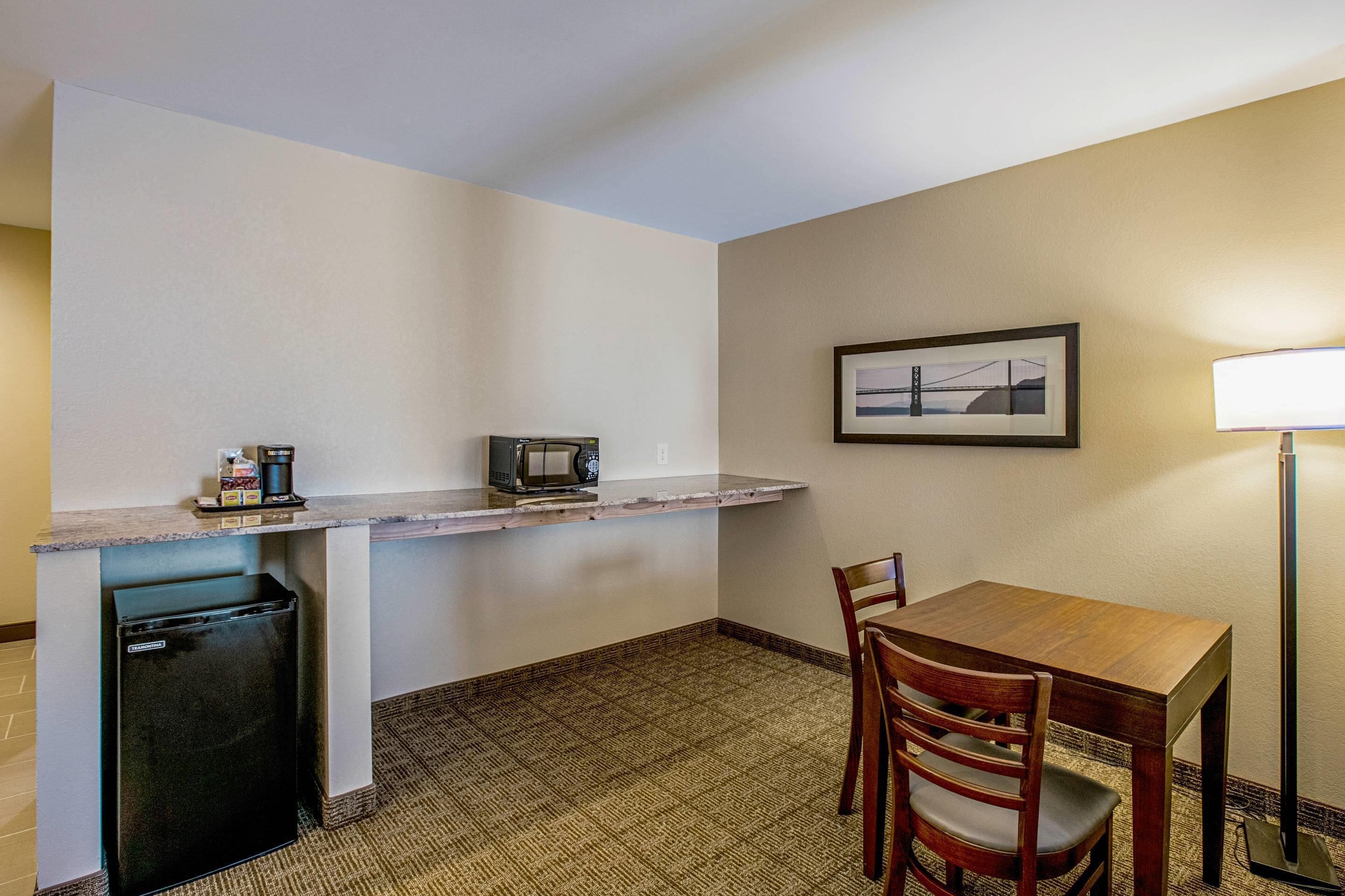 A compact kitchenette with a refrigerator, microwave, and coffee maker. There is a dining table with chairs and a framed picture on the wall.