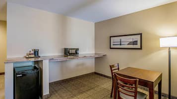 A compact kitchenette with a refrigerator, microwave, and coffee maker. There is a dining table with chairs and a framed picture on the wall.