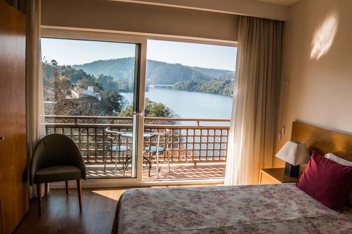 A hotel room with a bed, a chair, a nightstand, a lamp, and a balcony with a view of a lake and hills.