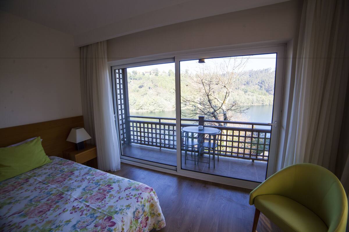 A hotel room with a bed, a chair, a table, and a balcony with a view of a lake and trees.