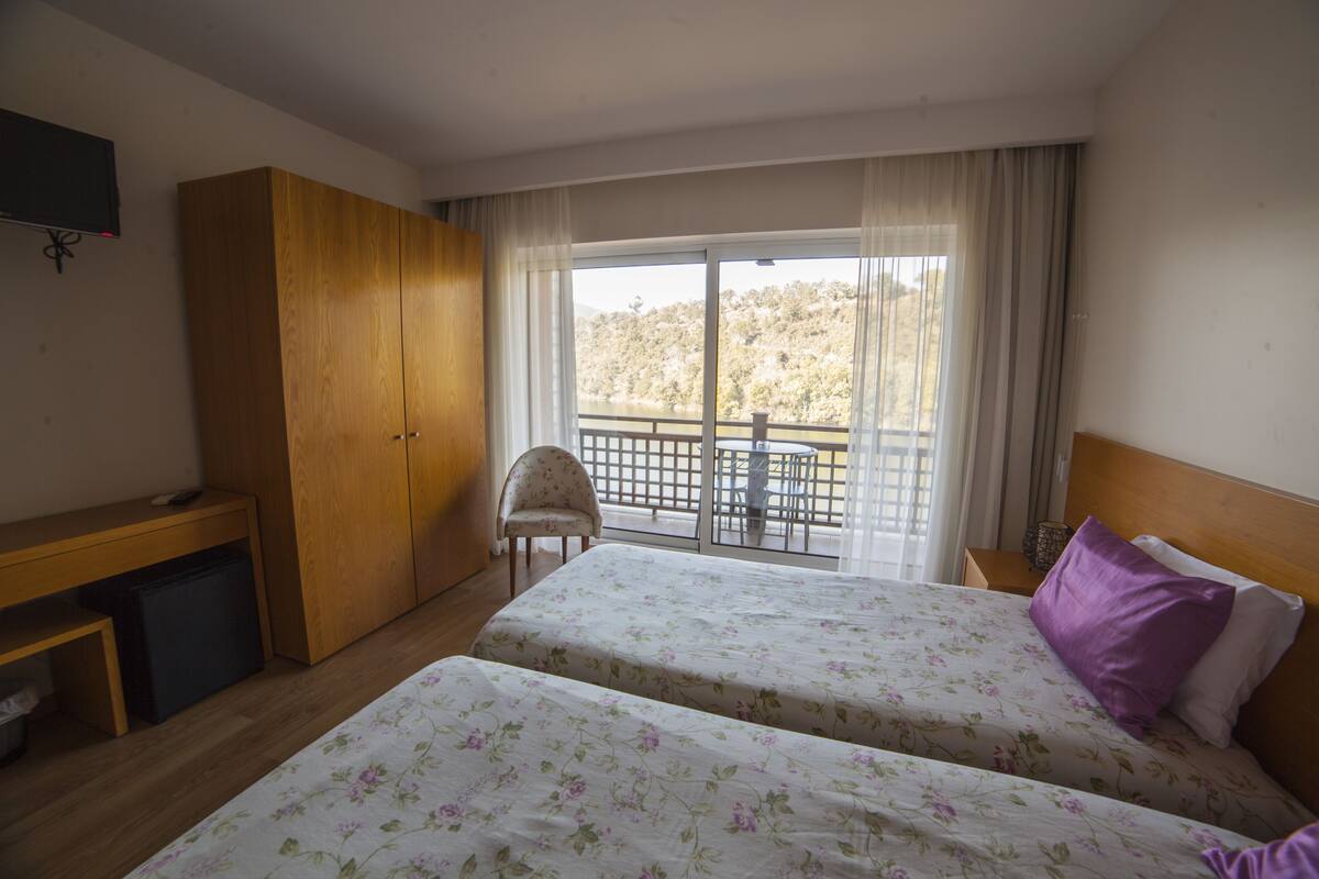 A hotel room with two beds, a desk, a chair, a TV, and a balcony with a view.