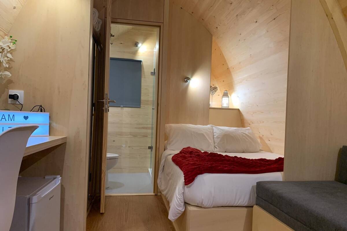 A compact room with a bed, bedside table, and a small desk with a computer. A bathroom is visible through an open door.