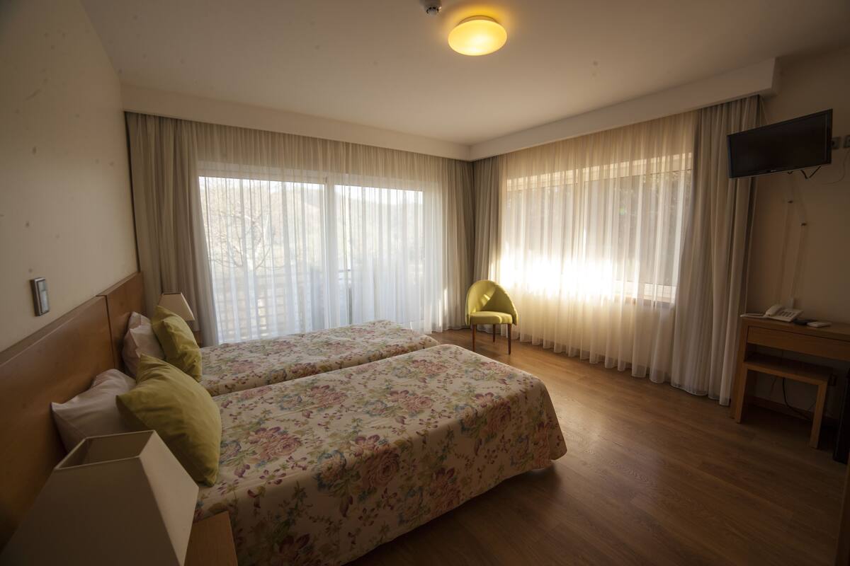 A hotel room with two beds, a desk, a chair, a TV, and large windows with curtains.