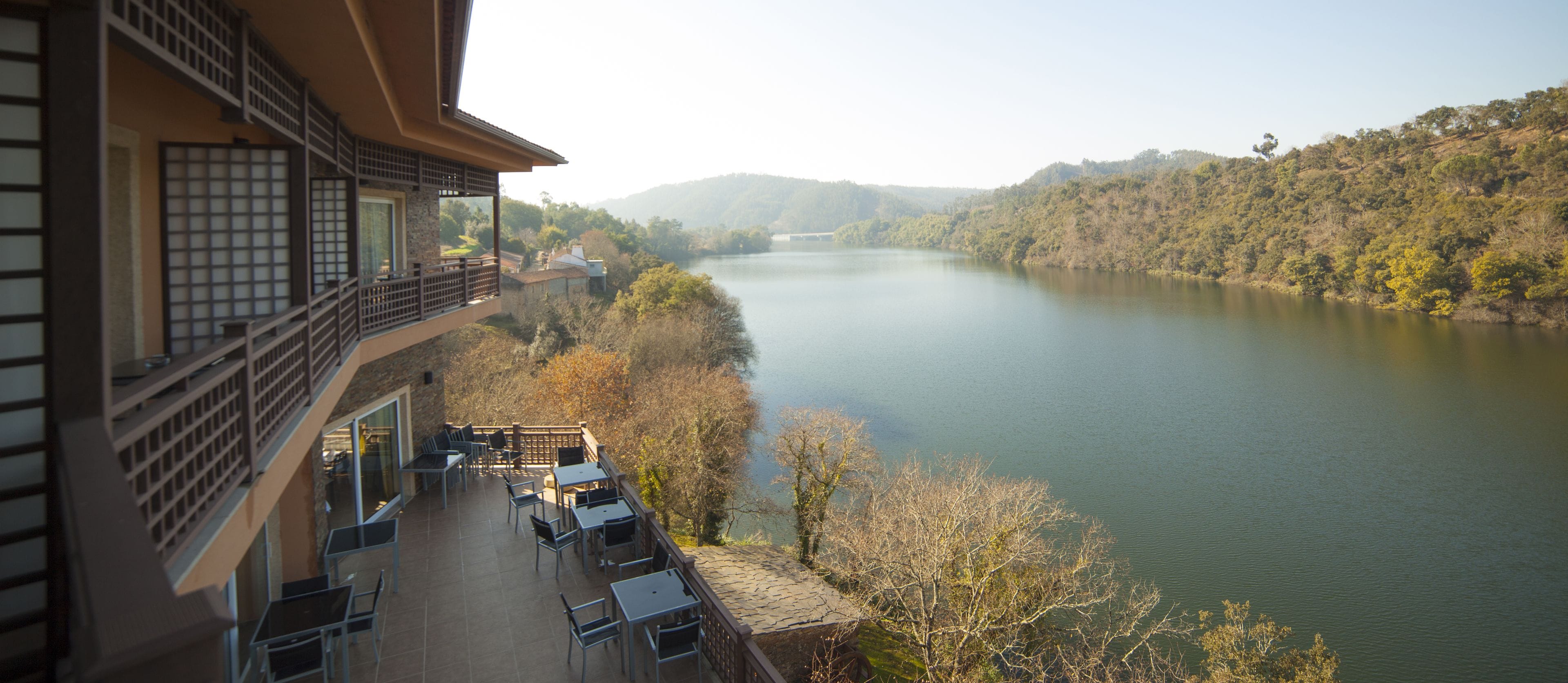 A balcony with outdoor seating overlooking a calm river and forested hills.