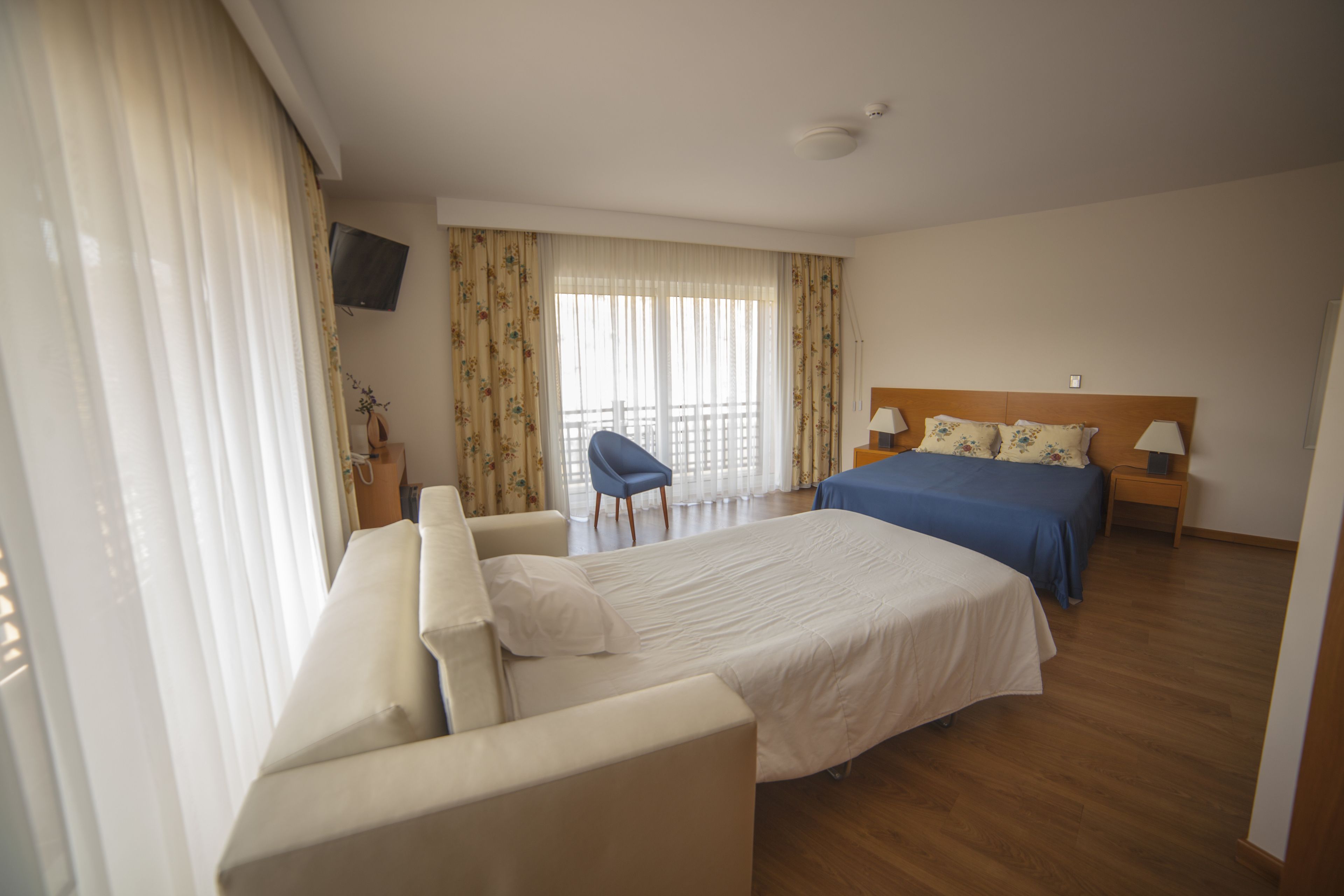 A hotel room with a large bed, a sofa, a chair, a TV mounted on the wall, and a balcony with curtains.