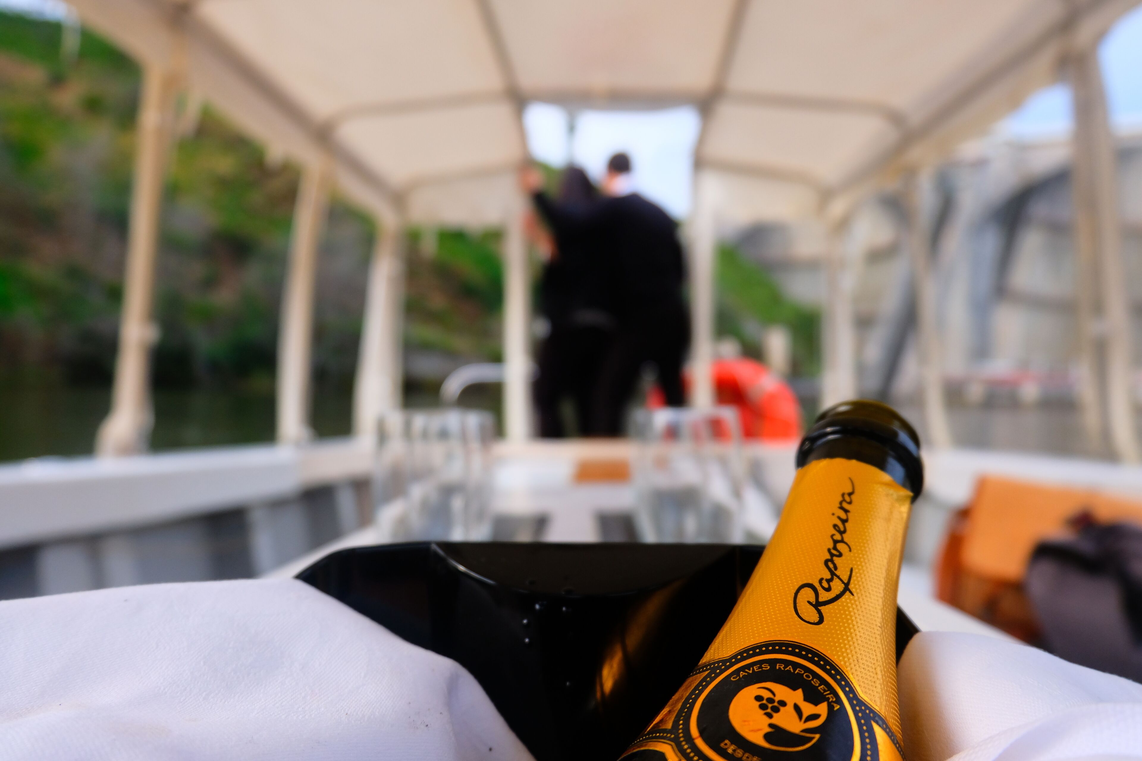 Una botella de champán en un barco con cubierta protegida y personas al fondo.