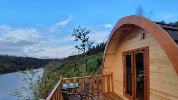 A wooden cabin with a curved roof, a deck with chairs, and a view of a river and hills. - Hotel Rural Quinta da Conchada (Penacova)