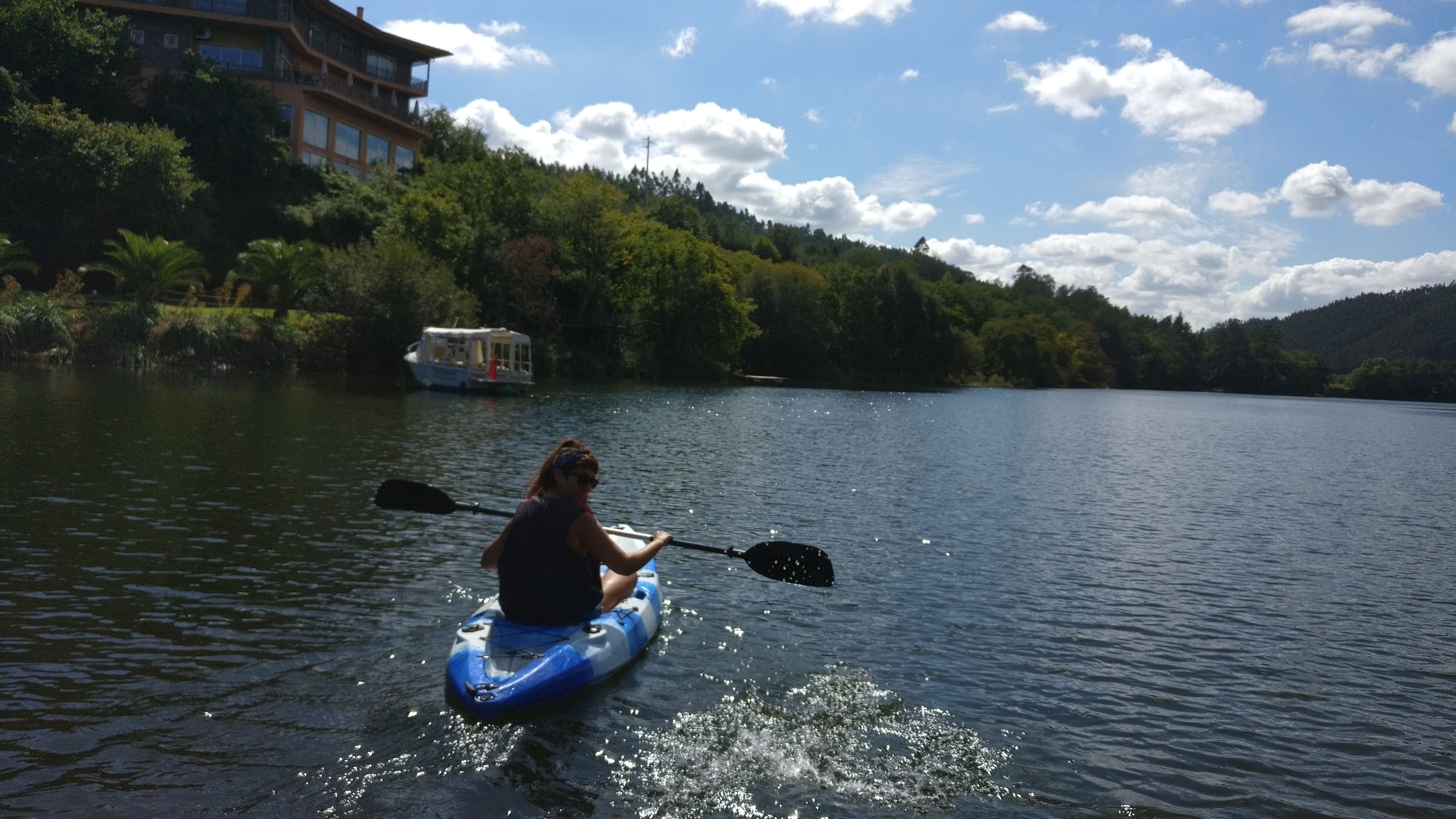 Une personne fait du kayak sur un lac calme, avec un bateau en arrière-plan et un bâtiment sur la rive.