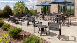 An outdoor patio with multiple tables and chairs, large umbrellas, and a brick building in the background. - Hampton Inn & Suites Overland Park South (Overland Park)