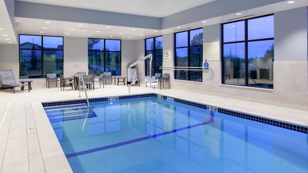 A modern indoor swimming pool with clear blue water, large windows, and a seating area with chairs and tables. - Hampton Inn & Suites Overland Park South (Overland Park)