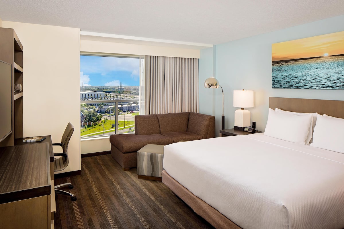A hotel room with a large bed, a desk, a chair, a sofa, a lamp, and a painting of a sunset over water.