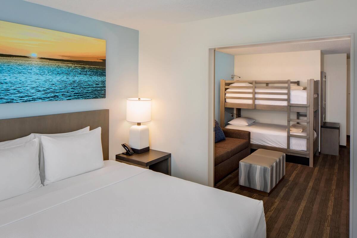 A hotel room with a bed, a nightstand with a lamp, a painting of a sunset over water, a bunk bed, and a small table.
