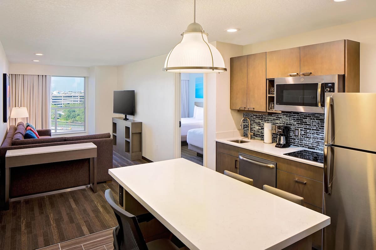 A modern hotel room with a kitchenette, a dining area, a sofa, a TV, and a window with a city view.