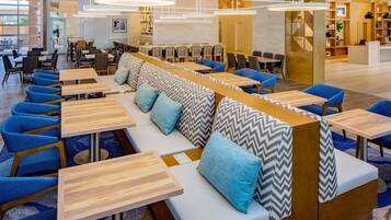 Ein modernes Restaurant mit langen Gemeinschaftstischen, blau gepolsterten Stühlen und einer zentralen Sitzgruppe mit gemusterten Kissen.