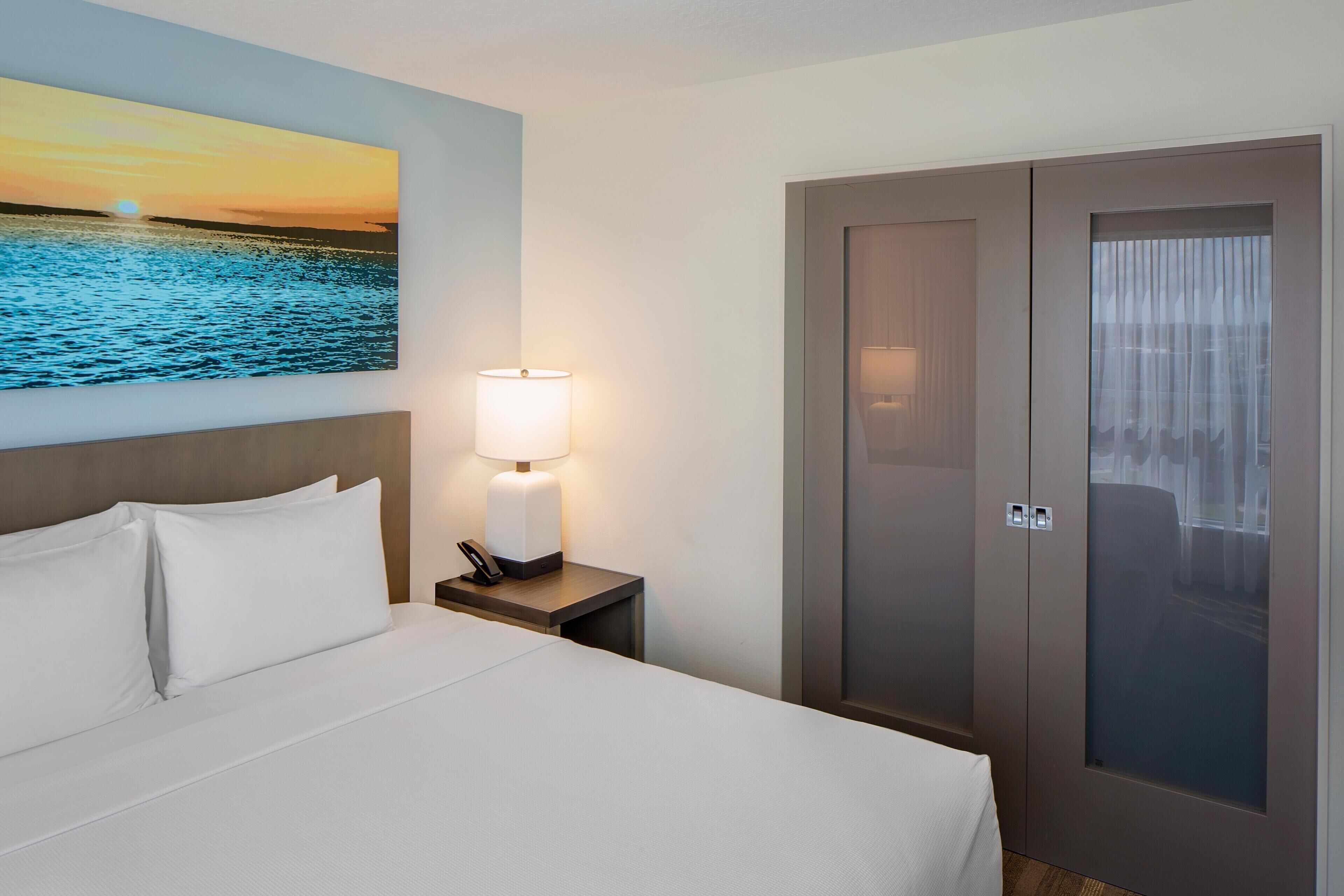 Une chambre d’hôtel avec un lit, une table de chevet avec une lampe, un tableau représentant un coucher de soleil sur l’eau et une armoire dont la porte est en verre dépoli.