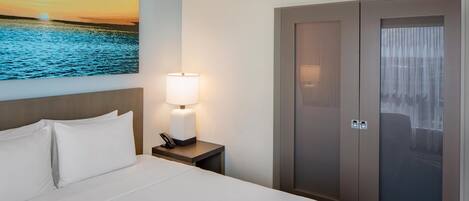 Une chambre d’hôtel avec un lit, une table de chevet avec une lampe, un tableau représentant un coucher de soleil sur l’eau et une armoire dont la porte est en verre dépoli.