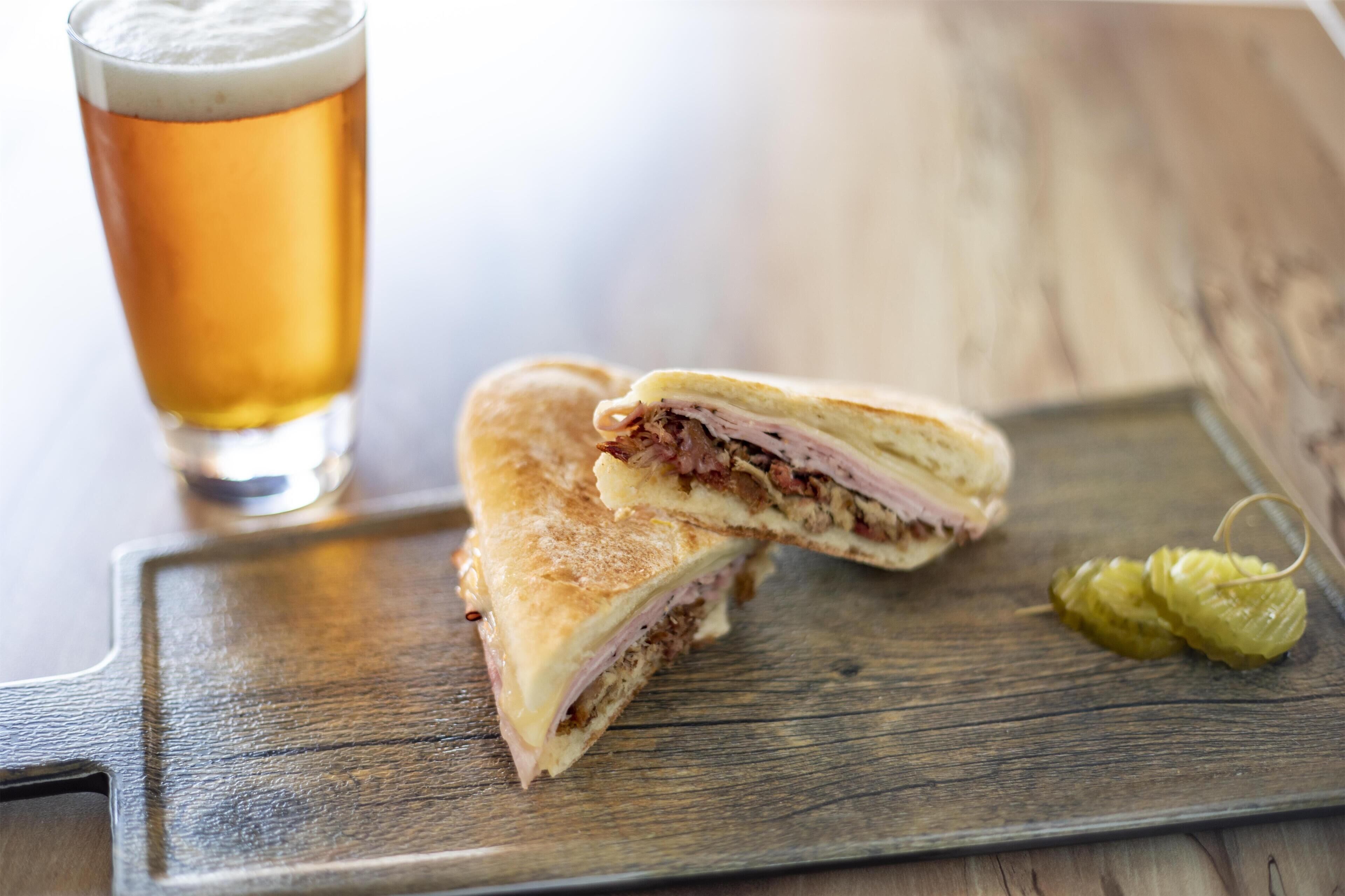 A sandwich cut in half on a wooden board, a glass of beer, and a pickle on the side.