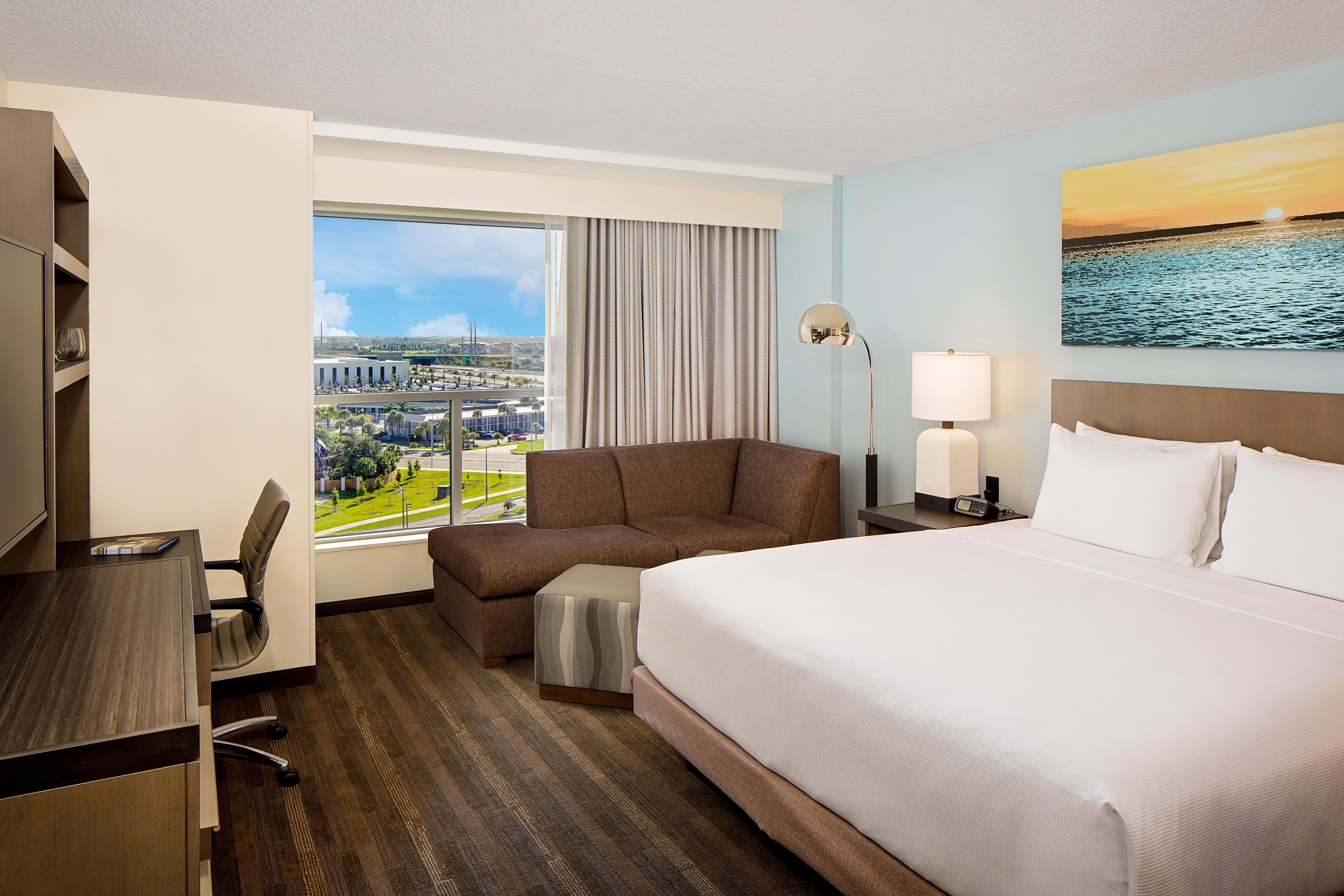 A hotel room with a large bed, a desk, a chair, a lamp, and a painting of a sunset over water.
