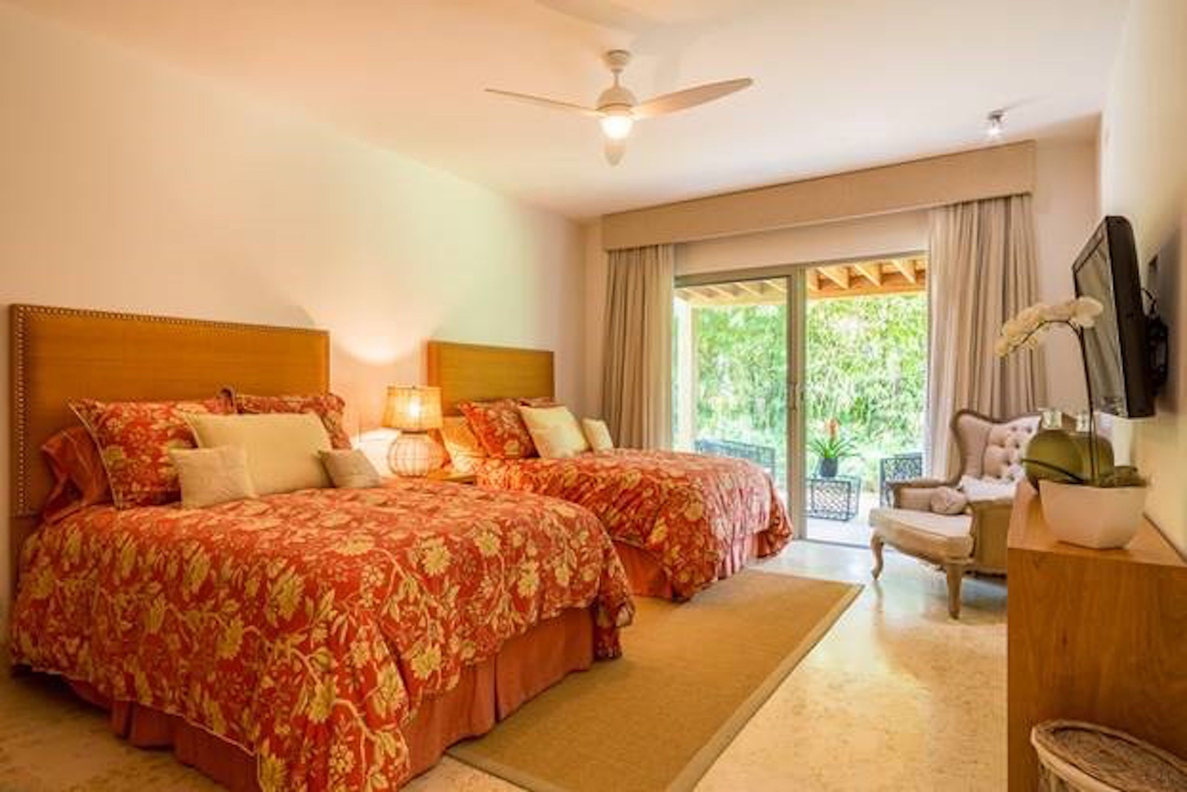 Habitación de hotel con dos camas, un ventilador de techo, un ventanal con cortinas y vistas al exterior con vegetación.