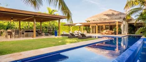 Piscina con sillones de descanso y una zona cubierta, rodeada de palmeras.
