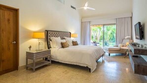 A bedroom with a bed, bedside tables, a ceiling fan, a TV, and a view of greenery outside.