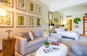 Una habitación de hotel moderna con un sofá gris, una mesa redonda con revistas, una cama con ropa de cama blanca y obras de arte con hojas enmarcadas en las paredes.