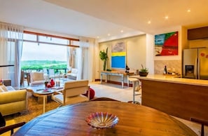 Un salon moderne avec une table à manger en bois, un canapé et un coin cuisine équipé d’un réfrigérateur, ainsi qu’un tableau accroché au mur.