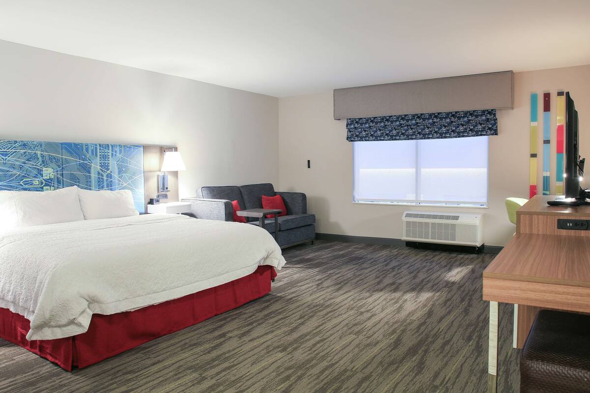 A hotel room with a large bed, a desk, a chair, a sofa, a window with a view, and a wall-mounted air conditioner.