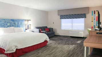 A hotel room with a large bed, a desk, a chair, a sofa, a window with a view, and a wall-mounted air conditioner.