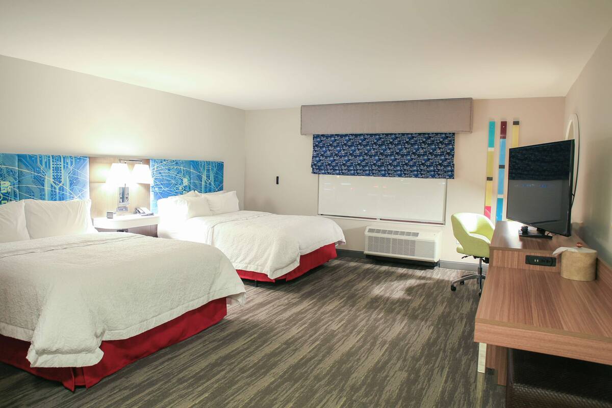 A hotel room with two beds, a desk, a chair, a TV, and a large window.